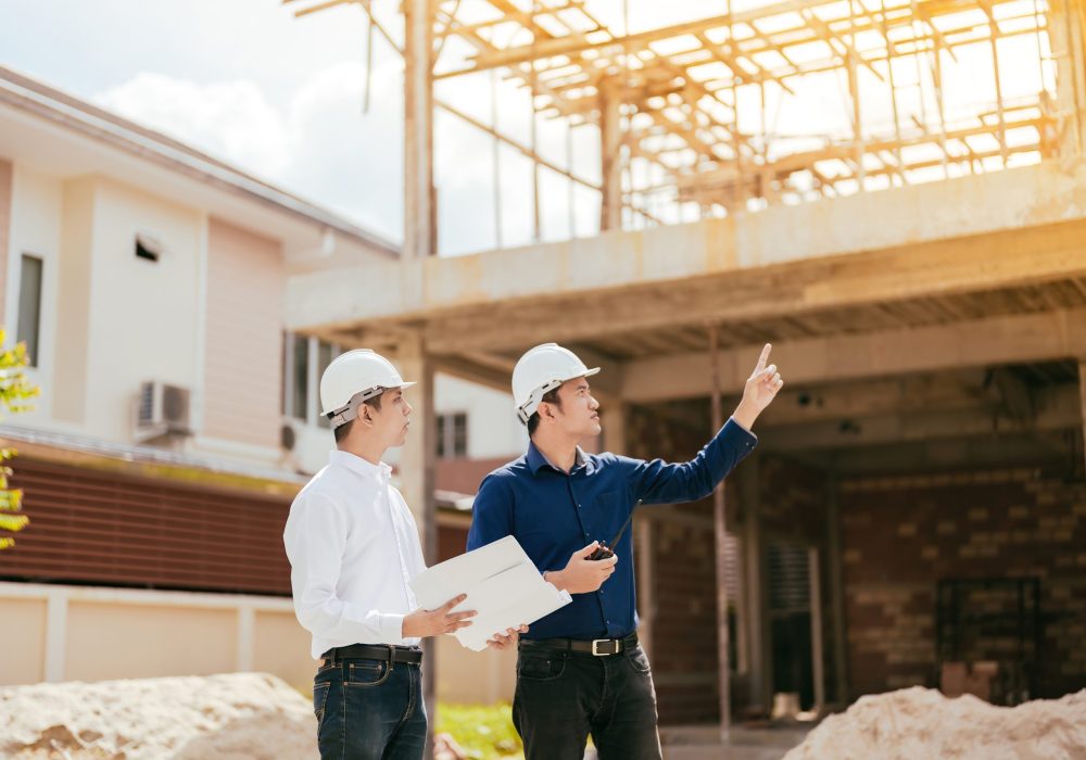 Two male engineers, Asians, are the technicians. Pointing to a newly built housing project related to the architectural industry, holding a blueprint, radio, wearing a hard hat and uniform.